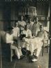 A photo of seven International Drillers around a table indoors with a white tablecloth. They are wearing white and four are sitting on wicker chairs. There is a jar, and a pipe, as well as a dog sitting on the table.