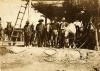 A photo of four men who are wearing hats standing with five horses, underneath a structure with a roof made from vegetation that has a wooden ladder leaning against it.