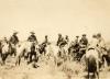 Une photo de dix hommes à cheval. Ils portent des chapeaux. Il y a de petits buissons au premier plan.