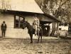 Une photo d'un homme en costume sur un cheval foncé. Il tient un fouet à la main. Il y a un bâtiment avec un revêtement et un porche couvert derrière lui. Un autre homme s'appuie contre le mur du bâtiment. 