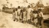 A photo of a group of children standing beside a dirt road. There is a building behind them and trees in the background.