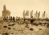 Un groupe d'hommes à cheval entourant un groupe de chevaux. Il y a des arbres à l'arrière-plan et une végétation basse au premier plan. 