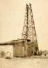 Four men standing in a dusty field beside a wooden oil rig with four sides. The bottom of the rig, with the machinery inside it, is covered in wooden boards and metal sheeting.