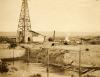 A photo of an oil rig behind a large dug hole with water in it and vertical posts. There is a pile of casing beside the rig with two men and a white horse standing beside it. There are hills in the background.