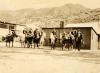 Une photo de neuf foreurs internationaux à cheval et portant des chapeaux. Deux bâtiments d'un étage se trouvent derrière eux et on aperçoit des collines vallonnées en arrière-plan.  