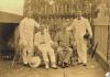 Two men sitting in chairs with a man standing on either side. They are wearing white and light coloured clothing. Each man is holding a hat or resting one on the ground. They are standing in front of a fence with vines on it. 