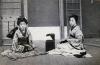 A photo of two girls dressed in Japanese clothing, sitting on folded mats. There is a dark box sitting open between them. There is a wall with matting behind them. 