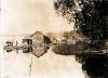 A photo of three small structures sided with reeds on a raft close to the shore of a river. There are people standing on the raft. There is a small boat to the left and reeds and a tree to the right. 