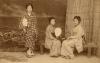 A photo of three girls holding fans, dressed in Japanese clothing. One is standing and the other two are sitting on a wooden bench. There is a reed mat against the wall and an illustrated scene of waves behind them. 