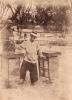 A man balancing a pole across his shoulders, with a wooden table on either side. A man is standing behind him. There is a picket fence and a couple of trees in the background. 