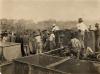 Bloss Sutherland and George York look on as a group of native workers stir cement in a metal barrel with sticks. There is another barrel beside them.  