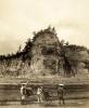 A photo of four people with two rickshaws standing on a road in front of a tall cliff with trees on the top of it. 