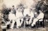 A photo of five International Drillers, wearing white, around a table with a hat on it. A servant stands behind them. There are trees in the background. 