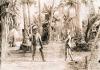 A photo of a man and a boy dressed in the traditional clothing of the Batak tribe. The man is holding a blade and the boy may be as well. They are standing in front of a number of trees.  