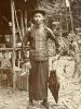 A photo of a Chinese man wearing a hat, buttoned shirt, and skirt. He is holding a closed umbrella at his side. There is a structure with wooden poles and a roof behind him.