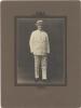 William McRae standing in a white suit and panama hat. Photograph is placed in a brown matte. 