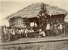 Group of local labourers living up to the left to receive payment from William McRae who is sitting at a desk in front of a hut.