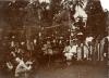 William McRae sitting in a white suit and pith helmet, surrounded by a group of locals under an awning. 