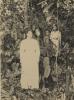 Mrs. McRae wearing a white dress, standing in the jungle with two native children in the background. 