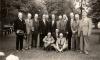 A group of retired International Drillers. Eleven are standing and three are sitting in front on grass. They are all wearing suits. There are trees behind them.