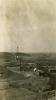 A photo of an oil derrick with four legs, surrounded by rolling hills. There are three small buildings in front of it with pipes running to the derrick.