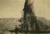 A photo of oil shooting over the top of a derrick with dark curtains around its base. There are dirt hills in the background.