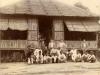 A group of people standing and sitting in front of a thatched bungalow. The doorway is open and there is a thatched window propped open on either side of the doorway.