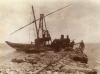 A photo of a boat in the water at the end of a stone pier. There is a crane lifting a steam boiler onto the rocks, and a man walks on the edge of the pier watching the boiler.