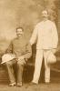 A photo of two International Drillers in front of a painted backdrop. On is sitting and one is standing. They are holding white pith helmets and are wearing  button-up shirts.