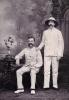 A photo of two men in front of a painted backdrop of trees. They are wearing white suits. There is a table beside the man sitting on the left, with books and flowers on it. The man on the right is wearing a white hat.