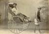 Une photo d'un homme assis dans un pousse-pousse à deux roues avec l'auvent refermé. Il est tiré par un homme de la région qui porte un chapeau de paille. Une toile de fond peinte se trouve derrière eux, avec des piliers et des arbres. 