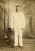 A photo of Thomas Knapp in front of a painted background of trees. He is wearing white and is resting his hand on the back of a chair with a hat on the seat.