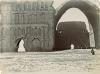 A photo of a group of people in front of a crumbling stone building. There is a giant archway on the left that rises three stories in the air. The wall on the left is carved with arches and pillars and there is a large triangular hole at the base.