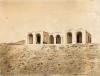 A photo of a concrete bungalow on a hill in the desert. There are two wings coming off of the centre part, each with two open arches. There are three arches in the middle section.