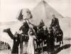 A photo of Mr. and Mrs. Saunders and their son riding camels. There are three local people standing on the ground and holding bottles on their heads. The Sphinx and a pyramid are in the background. 