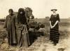 A photo of Annie Dyke standing beside a boy and woman holding a cow. Annie is wearing a dark striped dress, the woman a dark robe and head covering, and the boy a dark robe. There is a field behind them.