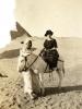 A photo of Annie Dyke in the desert, sitting on a white donkey that is being held by an Egyptian man. She is wearing a dark striped dress and hat, while he is in a white robe. There is a pyramid in the background.