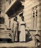 A photo of two men wearing long robes with dark hats and bands around their waists. They are standing beside a bench in front of a striped stone building.