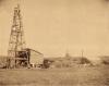 A photo of several International Drillers, wearing white suits and pith helmets, stand in front of an oil derrick in a grassy field. There is a small building and another oil derrick in the background.