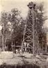 A photo of a group of International Drillers standing at the base and on the first support beam of an oil derrick. They are wearing white. There is a large cylinder and some logs in front of them and trees in the background.