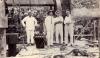 A photo of four men standing inside an oil derrick on a wooden plank floor. They are wearing white. There is a large wheel behind them and machinery beside them.