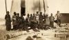 A photo of a crew of labourers with an International Driller who is wearing a pith helmet and standing on the right. They are in front of a derrick covered in cloth. There is a pile of casing in front of them.