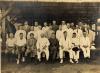 Group photograph of foreign drillers in Borneo all wearing white suits. They are sitting or kneeling on a raised platform underneath a roofed structure. 