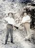 Two men in white shirts, hats, and dark pants. They are holding rifles. There is a small dog at the foot of the man on the right. They are standing in front of a wall of vegetation.