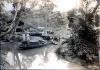 Six men on a boat in a narrow river. There are two men on a dock with a pile of casing beside them on the shore. A group of men stands behind the basing in front of three buildings. There are trees in the background.