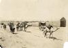 A group of camels with sacks on their backs, along with a few people, walking along a dirt road. There is a stone wall in the background.