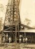 A group of men standing in front of a wooden oil rig. There is a ladder on the front side of the rig. The machinery beside the rig has a metal sheet roof over it.
