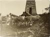 A photo of a group of workers in front of an oil rig covered in thatching. There are two oxen. One is attached to a cart with a piece of casing on it. There is a piece of casing hanging above the other ox, too.