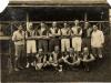 A photo of a group of men wearing matching shirts with black on one side and light on the other. Eight are standing with their arms crossed. Four are sitting cross-legged with a ball on a stand in front of them.