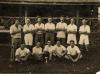 A photo of a group of men wearing white shirts. Seven are standing with their arms crossed. Five are sitting cross-legged with a ball on a stand in front of them.